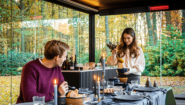 Par som dekker bord under pergola med varmelist, høstskog utenfor glasset